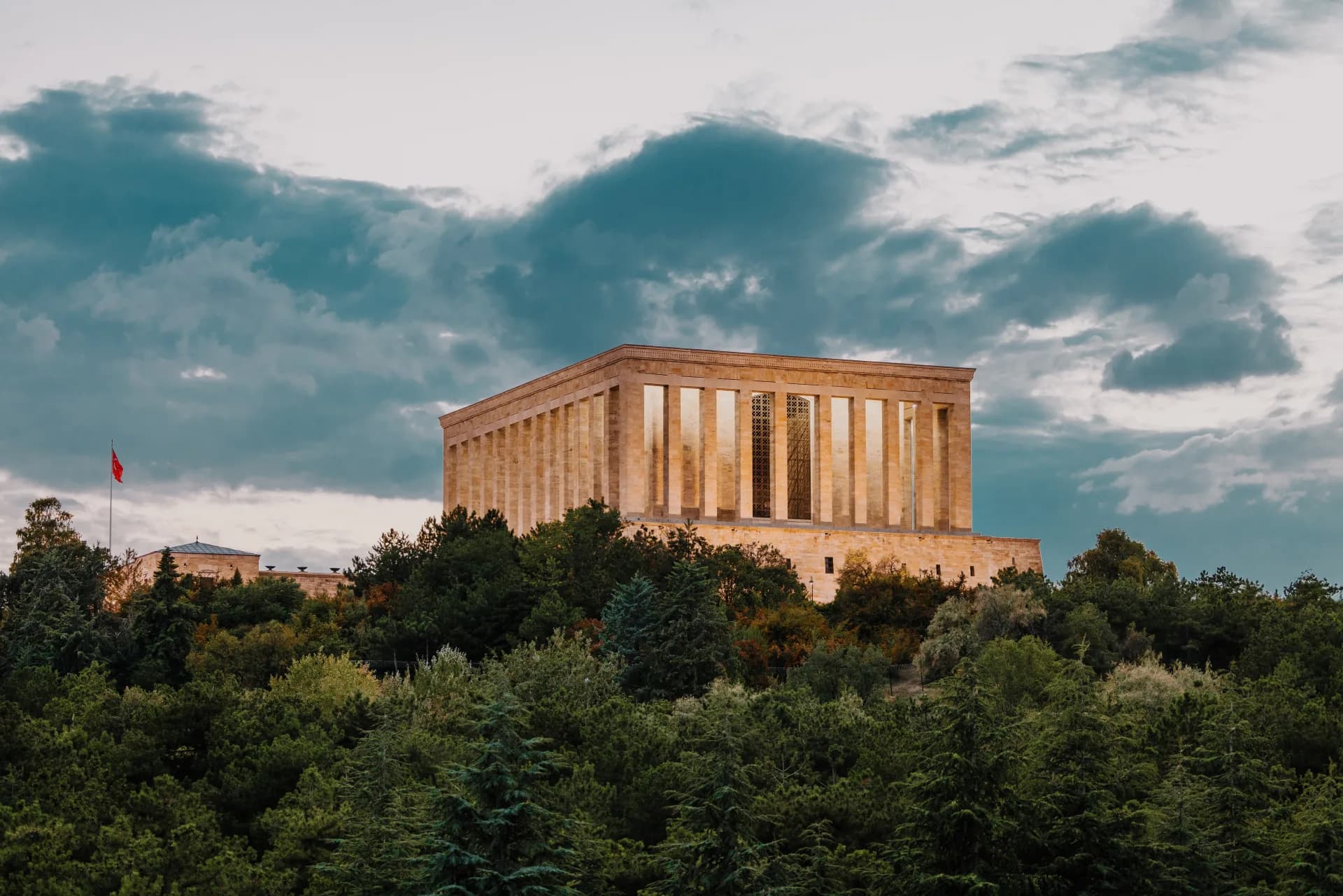 Anıtkabir, ağaçlık tepenin üzerinde; solda Türk bayrağı, gökyüzü bulutlu